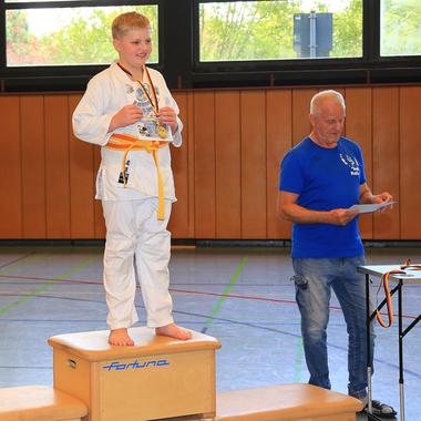 Junge mit Medaille steht auf einem Podest, während ein Mann neben ihm ein Dokument liest. Tisch mit Pokalen im Hintergrund.