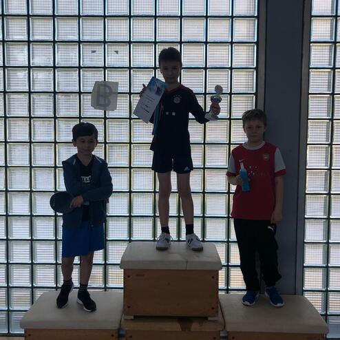 Drei Kinder stehen auf Podesten, der mittlere hält einen Pokal und eine Urkunde, während die anderen einen Tischtennisschläger und Getränke halten.