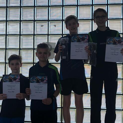 Vier Jungen stehen in einer Reihe, halten Urkunden und Preise vor einer Glaswand. Licht strömt durch die Platten.