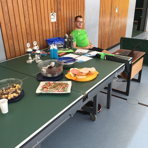 Mann in grünem Shirt sitzt an einem Tisch mit Tischtennismaterial und Snacks, vor einer holzvertäfelten Wand.