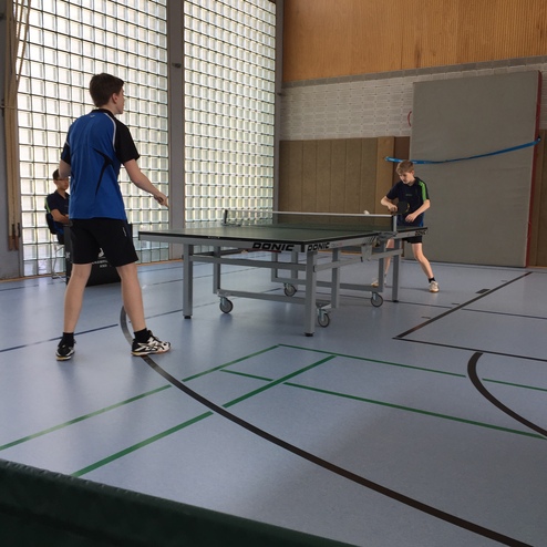 Zwei Jungen spielen Tischtennis an einem Tisch in einer hellen Sporthalle mit großen Fenstern.