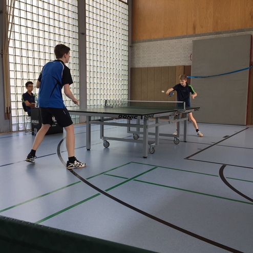 Zwei Jungen spielen Tischtennis an einem Tisch, während ein weiterer Junge zuschaut. Sporthalle mit großen Fenstern.