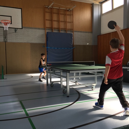 Zwei Jungen spielen Tischtennis in einer Sporthalle, einer mit Schläger bereit zum Schlag, der andere in Position zum Empfang.