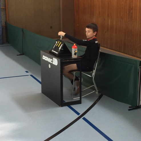 Junge sitzt an einem Tisch mit Tischtennis-Equipment und einer Wasserflasche in einer Sporthalle.