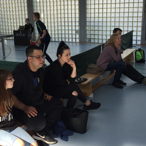 Vier Personen sitzen auf einer Bank in einem Sportzentrum, während ein Tischtennisspiel im Hintergrund stattfindet.