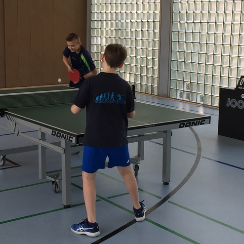 Zwei Kinder spielen Tischtennis an einem Tisch, während ein dritter Junge das Spiel zählt.