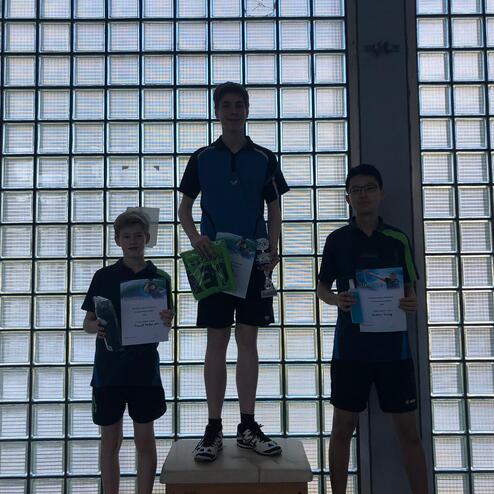 Drei Tischtennisspieler stehen auf einem Podest mit Urkunden und Pokalen vor einer Glaswand.