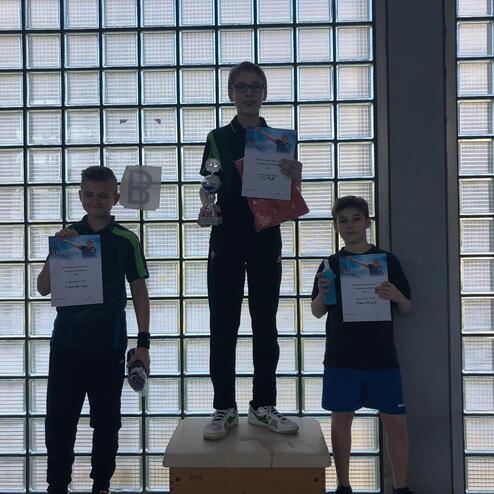 Drei Kinder stehen auf einem Podest, halten Urkunden und einen Pokal, vor einer Wand aus Glasbausteinen.