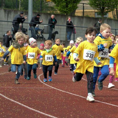 Gruppe von Kinder im gelben T-Shirts beim Laufen auf einer Laufbahn während eines Wettkampfs. Zuschauer im Hintergrund.