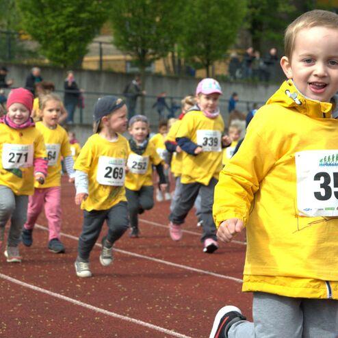 Gruppe von Kindern in gelben T-Shirts läuft auf einer Laufbahn bei einem Wettkampf, einige mit Startnummern.