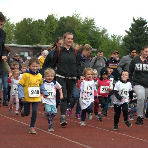 Gruppierte Kinder in Laufshirt rennen auf einer Laufbahn, begleitet von Erwachsenen, während sie an einem Wettlauf teilnehmen.