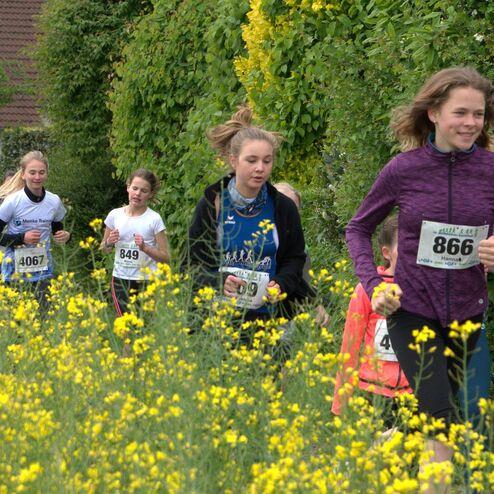 Gruppe von Mädchen joggt in Laufkleidung durch blühendes, gelbes Feld entlang eines grünen Weges.