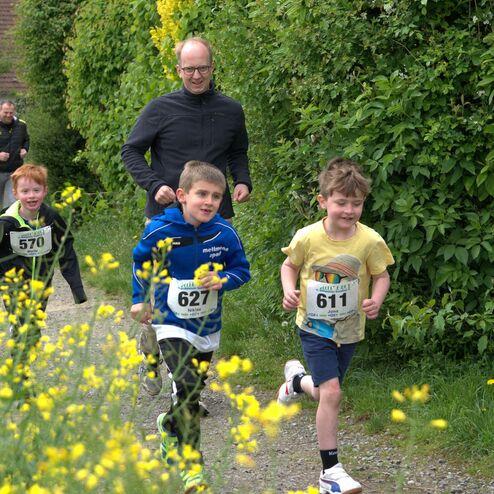 Gruppe von Kindern mit Startnummern, die fröhlich einen schmalen Weg zwischen Blumen entlang laufen.