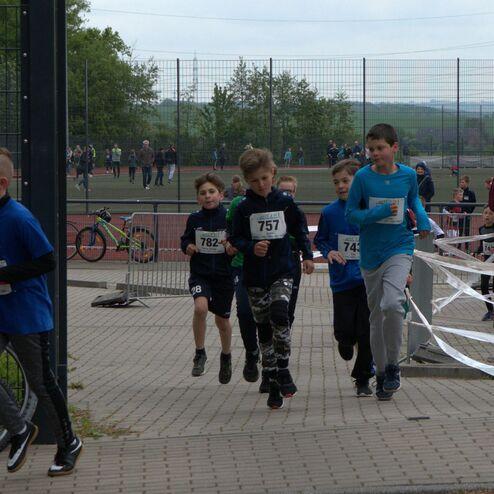 Sechs Kinder in Sportkleidung laufen bei einem Laufwettbewerb, im Hintergrund sind Zuschauer und Fahrräder zu sehen.
