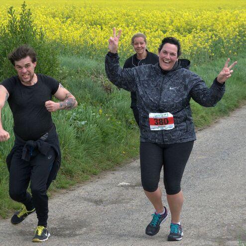 Drei Läufer auf einem Weg, umgeben von gelben Rapsfeldern, einer lächelt und zeigt Victory-Zeichen.