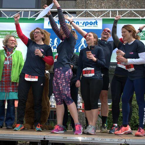 Gruppe von Sportlerinnen feiert auf der Bühne mit Urkunden und Tassen in der Hand nach einem Wettkampf.
