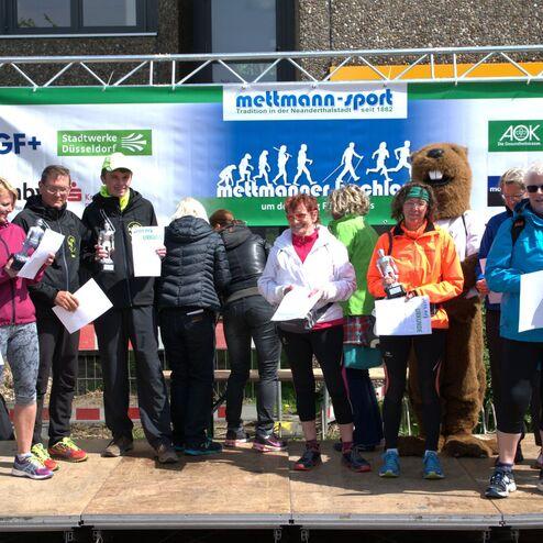 Gruppenfoto von Teilnehmern einer Laufveranstaltung mit Pokalen und Urkunden auf einer Bühne.
