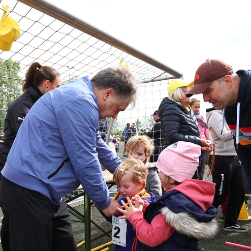 Eine Gruppe von Erwachsenen und Kindern steht um einen Tisch am Sportplatz, während ein Mann einem Kind etwas erklärt.