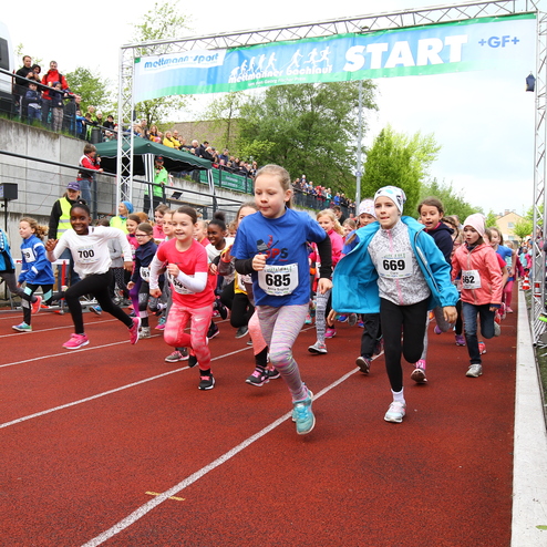Viele Kinder starten zu einem Laufwettbewerb auf einer roten Laufbahn, Zuschauer beobachten vom Rand.
