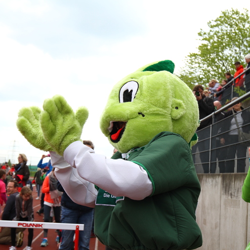 Grüner Maskottchen applaudiert auf einem Sportplatz, umgeben von Zuschauern und Kindern am Wettkampfort.