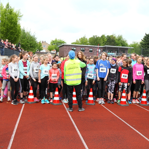 Gruppe junger Läuferinnen steht an der Startlinie eines Sportevents, beobachtet von Zuschauern auf der Tribüne.