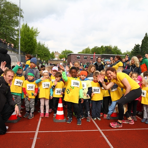 Gruppe von Kindern in gelben T-Shirts steht auf einer Laufbahn, einige winken, hinter ihnen Zuschauer und eine grüne Maskottchenfigur.