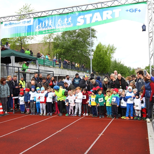 Startlinie eines Kinderlaufs mit Teilnehmern in bunten Trikots, umgeben von Zuschauern auf einer Sportanlage.