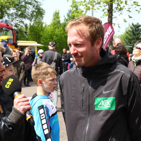 Interview-Szene mit einer Frau, die mit einem Mann in Sportkleidung spricht, während im Hintergrund Zuschauer stehen.