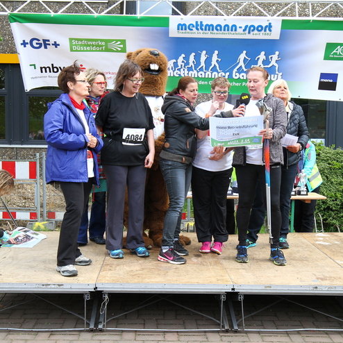 Gruppe von fünf Personen auf einer Bühne hält eine Preistransparente, während ein Maskottchen im Hintergrund steht.