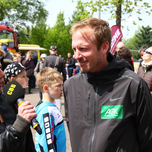 Interview mit einer Frau mit Mikrofon und einem Mann in Jacke, umgeben von Zuschauern bei einer Veranstaltung im Freien.