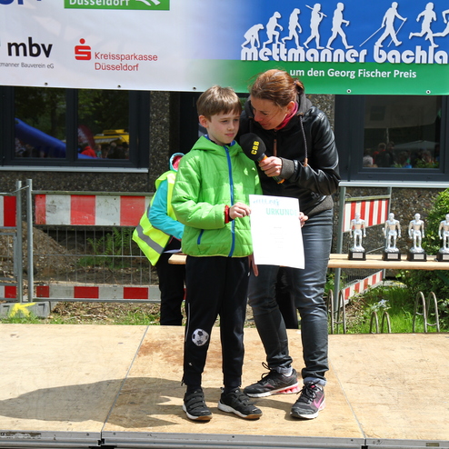 Junger Junge in grüner Jacke erhält eine Urkunde von einer Frau auf einer Bühne, im Hintergrund stehen Pokale.