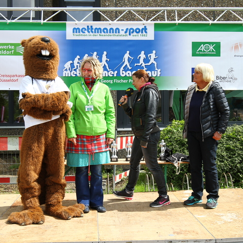 Veranstaltungsbühne mit einem Maskottchen, drei Personen und einem Banner im Hintergrund. Trophäen sind sichtbar.