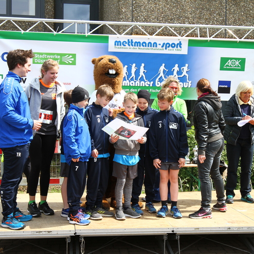 Gruppe von Kindern und Erwachsenen auf einer Bühne, die den Preis bei einem Sportevent entgegennehmen, umgeben von Sponsorenbanner.