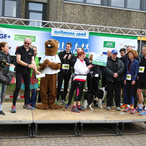 Sieben Personen und ein Maskottchen stehen auf einer Bühne bei einer Veranstaltung, während eine Sprecherin das Mikrofon hält.