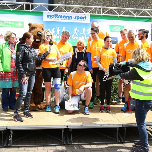 Gruppenshow mit sportlich gekleideten Personen in orange T-Shirts auf einer Bühne, während eine Frau spricht.