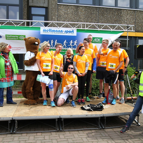 Gruppe von Läufern in orangefarbener Sportbekleidung posiert auf einer Bühne mit Maskottchen und Veranstaltern.