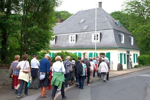 Gruppe von Menschen, die an einem Haus mit grünen Fensterläden entlanggeht, umgeben von Bäumen und Sträuchern.