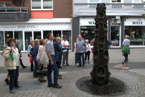 Gruppe von Menschen betrachtet eine hohe Skulptur auf einem Pflasterweg in einer Fußgängerzone.