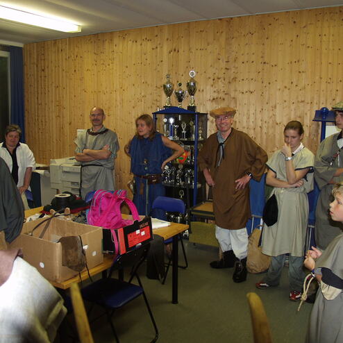 Gruppenszene in einem Raum mit mehreren Personen in historischen Kostümen und einem Tisch voller Requisiten.