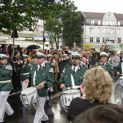 Schüler und Musiker in grüner Uniform marschieren bei einer Parade mit Trommeln und Pfeifen durch eine belebte Straße.