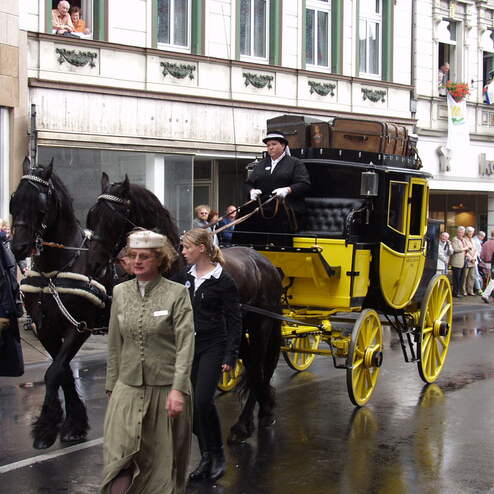 Pferdegepäckwagen mit schwarzem und gelbem Kasten, gezogen von zwei Pferden, in einer belebten Stadtstraße.