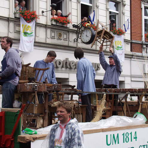 Teilnehmer einer Parade stehen auf einem Wagen, umgeben von Stühlen und Zuschauern, die aus Fenstern winken.
