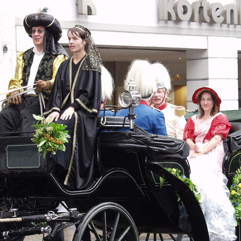 Fünf Personen in historischen Kostümen sitzen in einer befestigten Pferdekutsche vor einem modernen Gebäude.