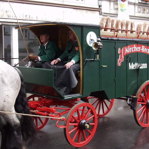 Pferdezugwagen mit Brot, besetzt von zwei Männern in grünen Jacken, fährt durch eine belebte Straßenlandschaft.