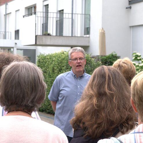 Mann spricht zu einer Gruppe Menschen vor modernen Gebäuden mit Büschen und Balkonflächen im Hintergrund.