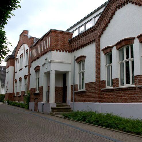 Historisches Backsteingebäude mit weißen Fassaden und großen Fenstern an einer gepflasterten Allee.