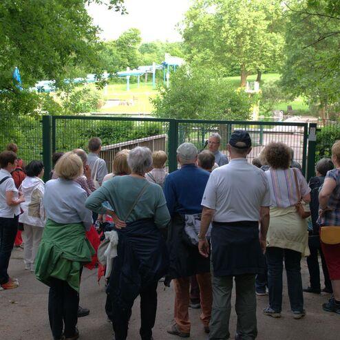 Gruppe von Menschen schaut durch ein Tor auf ein Freizeitgelände mit grüner Wiese und Rutschen im Hintergrund.