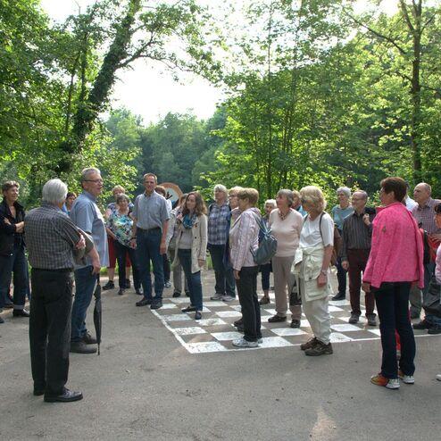Gruppe von Menschen steht auf einem Parkplatz im Wald, während jemand ihnen anspricht. Umgeben von Bäumen.