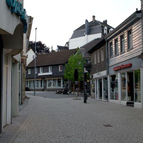 Leere Einkaufsstraße mit modernen Gebäuden, gepflastertem Weg und Bäumen, im Hintergrund eine ruhige Ecke mit Café.