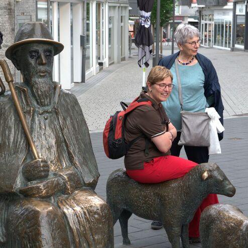 Skulptur eines alten Hirten mit einem Stock, während eine Frau auf einem Schaf sitzt und zwei weitere Frauen stehen daneben.